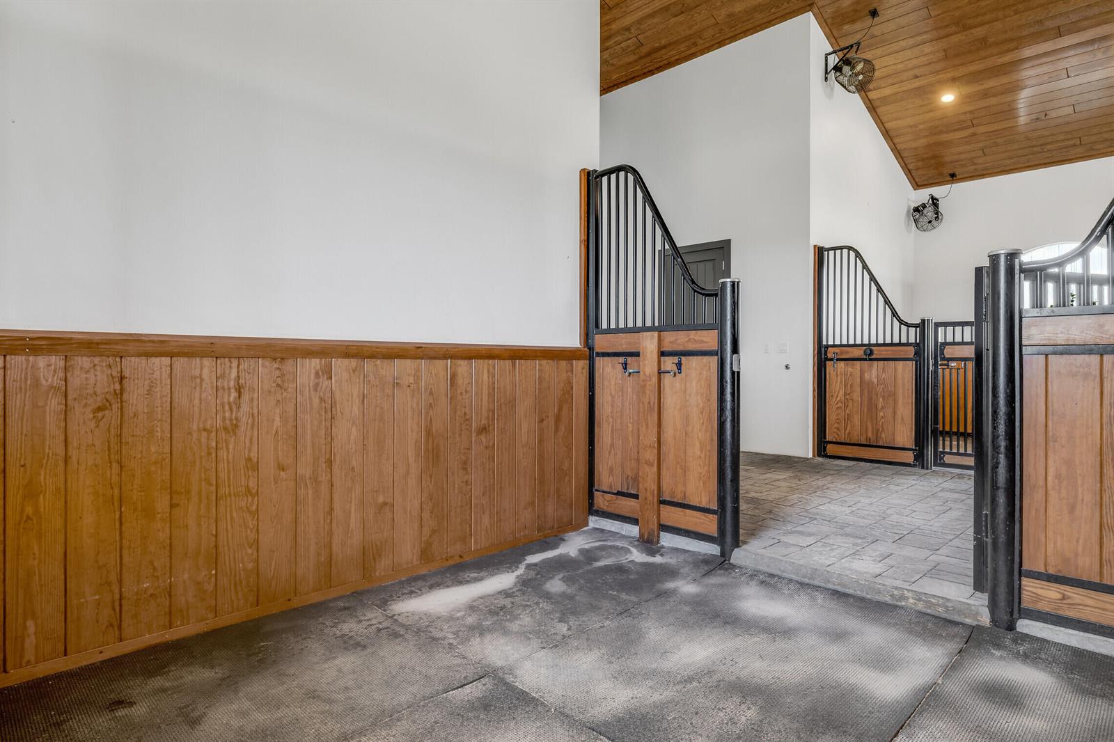 A horse stable aisle features wood-paneled walls, stall doors with black metal grilles, and flooring — 5181 Lasso Way, Wellington FL 33449, WELLINGTON, FL