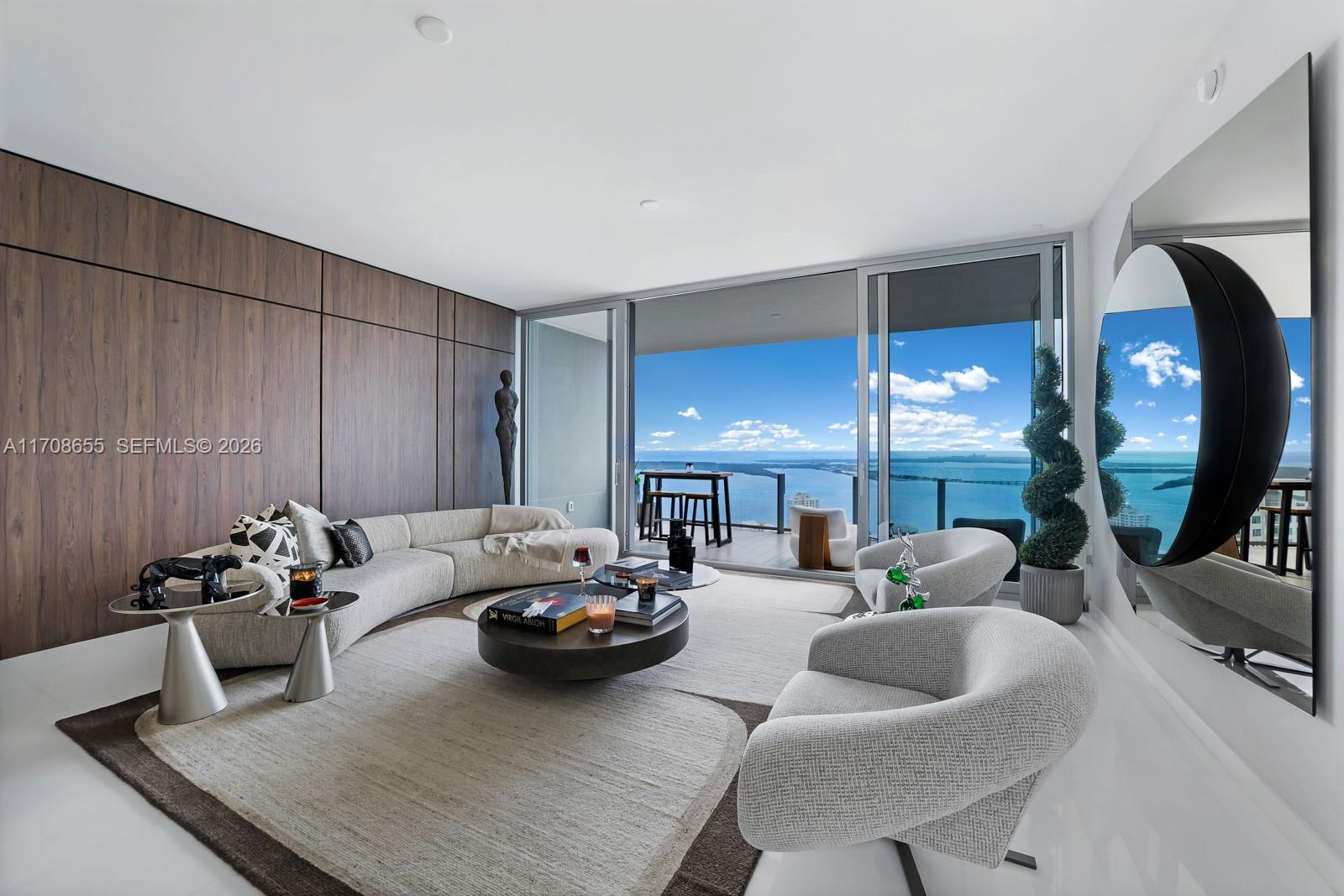 A modern living room features a curved sofa, wood-paneled wall, and floor-to-ceiling windows overlooking water — 300 Biscayne Blvd Way # 4703, Miami FL 33131, MIAMI, FL