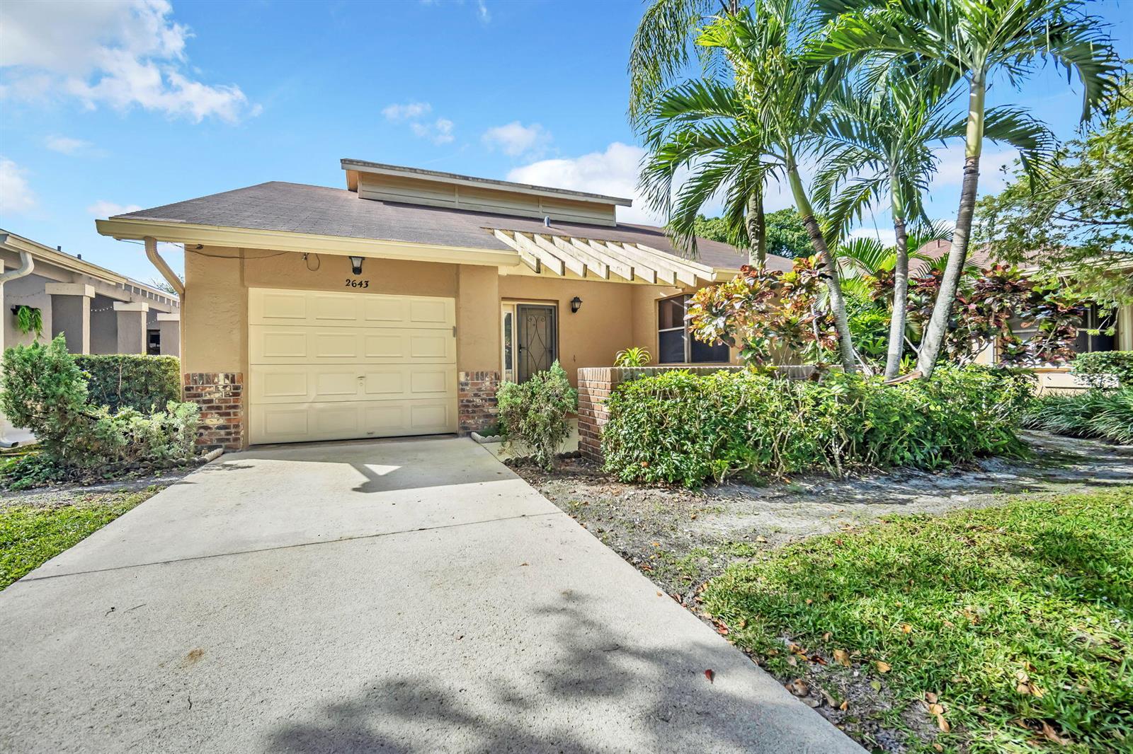 A tan single-story house features a beige garage door, a wooden pergola, and palm trees — 2643 Calliandra Terrace, Coconut Creek FL 33063, COCONUT CREEK, FL