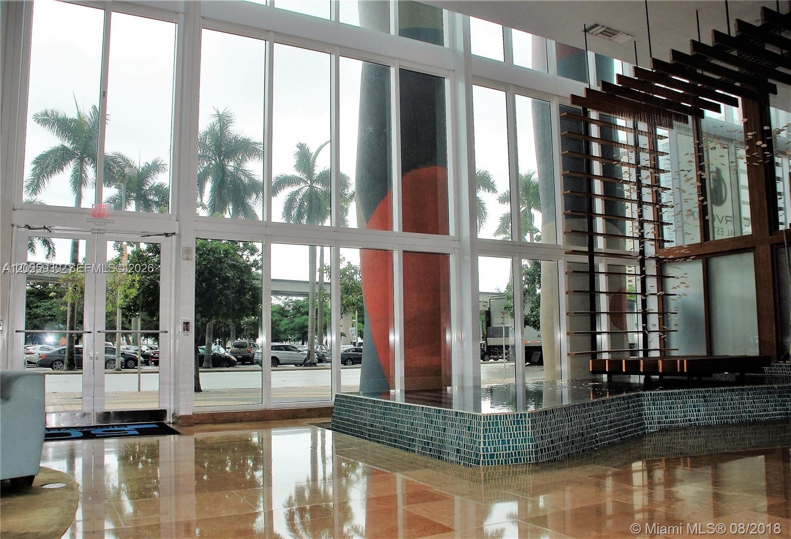 A modern lobby features floor-to-ceiling windows, a decorative water feature, and a large structural column — 50 Biscayne Blvd # 2105, Miami FL 33132, MIAMI, FL