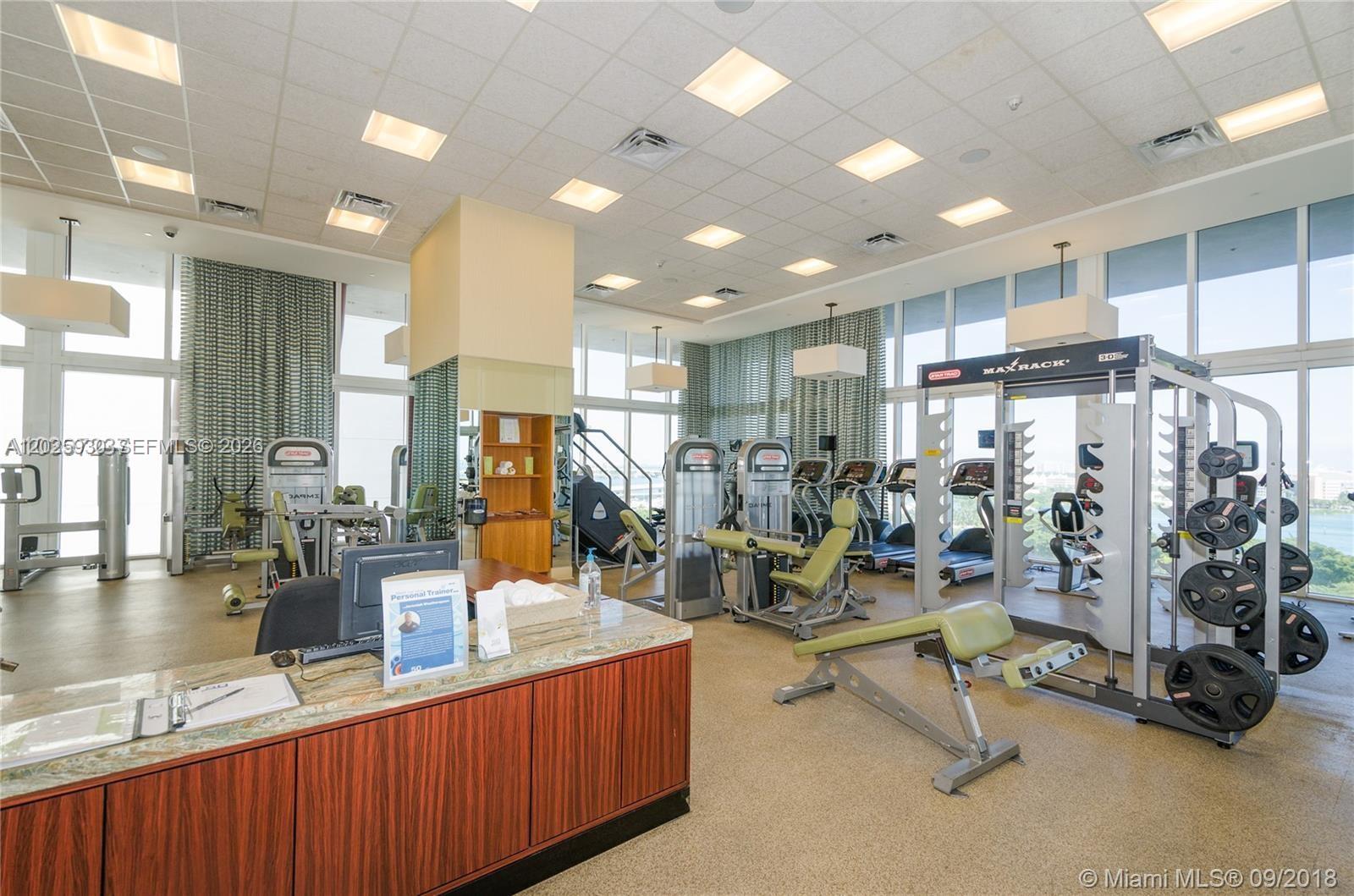 A fitness center features gym equipment, a reception desk, and large windows with curtains — 50 Biscayne Blvd # 2105, Miami FL 33132, MIAMI, FL