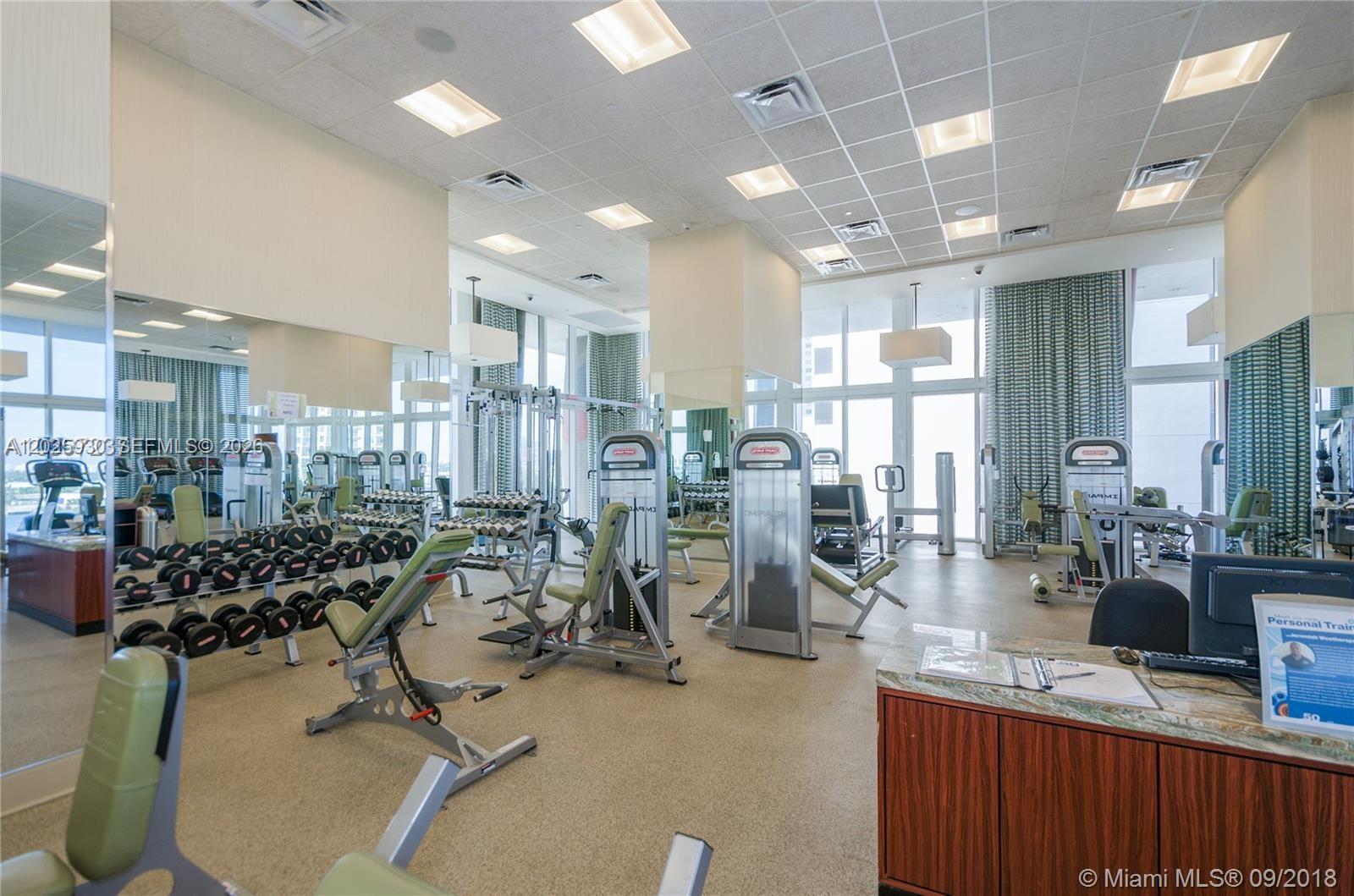 A fitness center features weight machines, dumbbell racks, mirrored walls, and a reception desk — 50 Biscayne Blvd # 2105, Miami FL 33132, MIAMI, FL