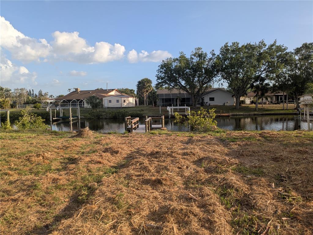 A grassy lot overlooks a canal with wooden docks and houses in the background — 18641 KLINGLER CIRCLE, PORT CHARLOTTE, FL