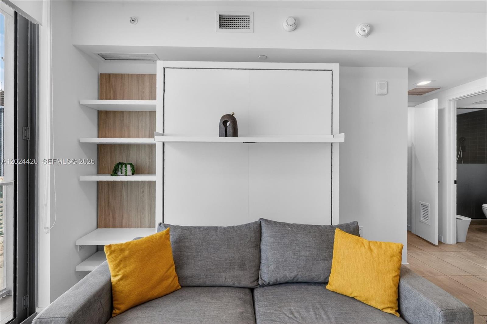 A gray sofa sits in front of a white wall-mounted Murphy bed and shelving unit — 227 NE 2nd St # 2805, Miami FL 33132, MIAMI, FL