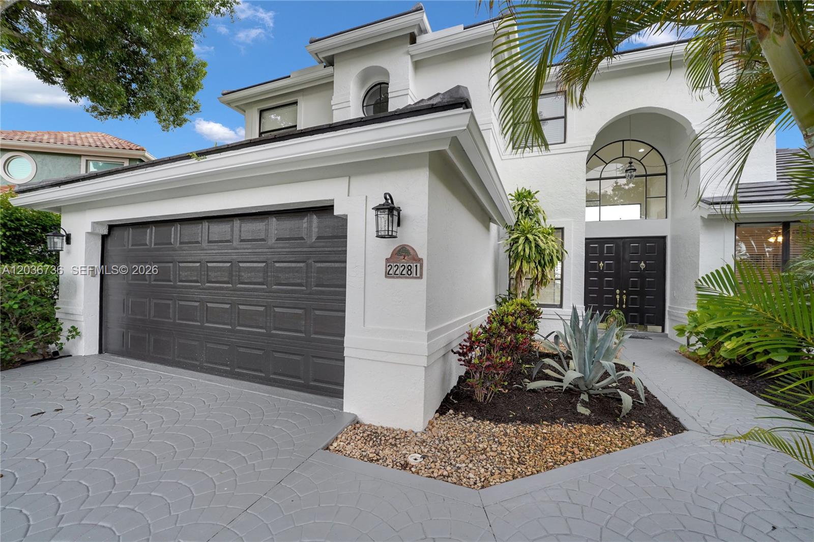A white two-story house features a dark garage door, arched entryway, and patterned driveway — 22281 Tupelo Pl, Boca Raton FL 33428, BOCA RATON, FL