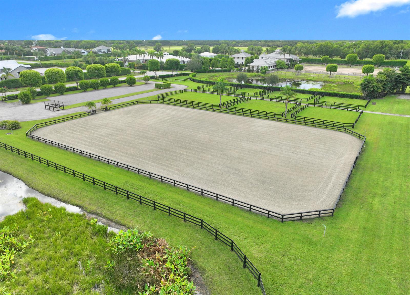 An aerial view shows a large, fenced equestrian riding arena surrounded by green grass — 3899 Shutterfly Way, Wellington FL 33414, WELLINGTON, FL