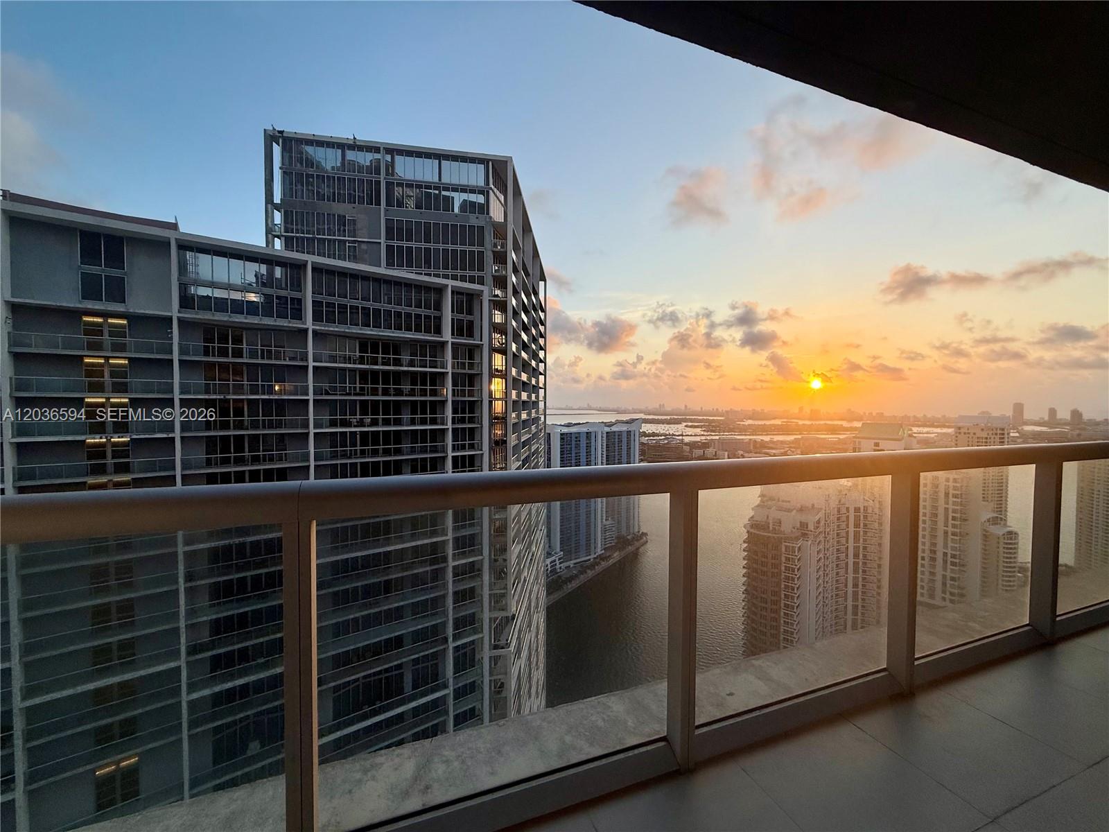 A balcony view overlooks a modern high-rise building and a sunset over the water — 495 Brickell Ave # 5106, Miami FL 33131, MIAMI, FL