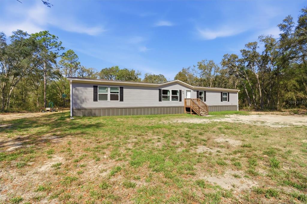 A single-story manufactured home with gray siding sits on a large, grassy, wooded lot — 7290 NE 110TH AVENUE, BRONSON, FL