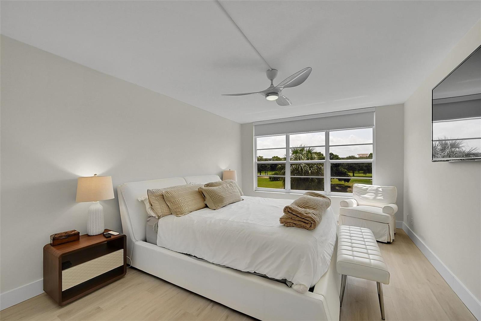 A bedroom features a white bed, a wooden nightstand, a ceiling fan, and a window — 555 Oaks Lane # 311, Pompano Beach FL 33069, POMPANO BEACH, FL