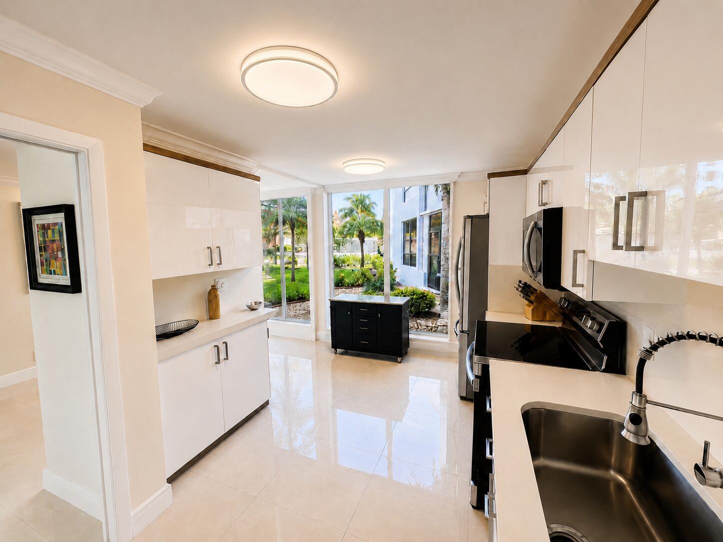 A modern kitchen features white glossy cabinets, stainless steel appliances, and large windows overlooking greenery — 7170 PROMENADE Drive # 101, Boca Raton FL 33433, BOCA RATON, FL