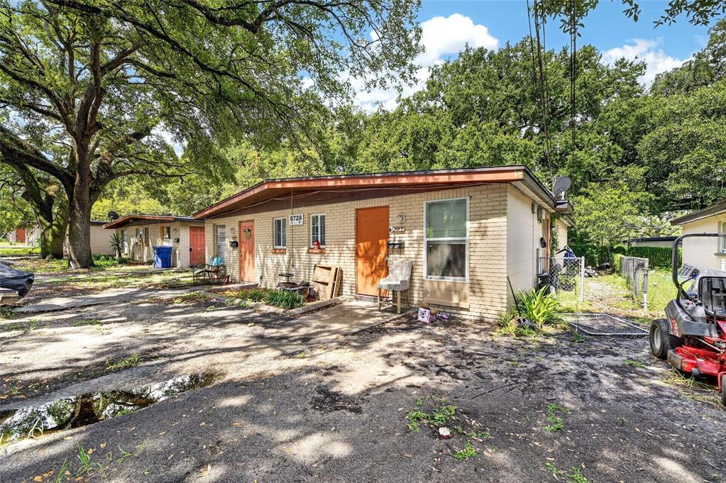 A single-story brick duplex with orange doors sits on a dirt lot under large trees — 8726 & 8728 N 48TH STREET, TAMPA, FL