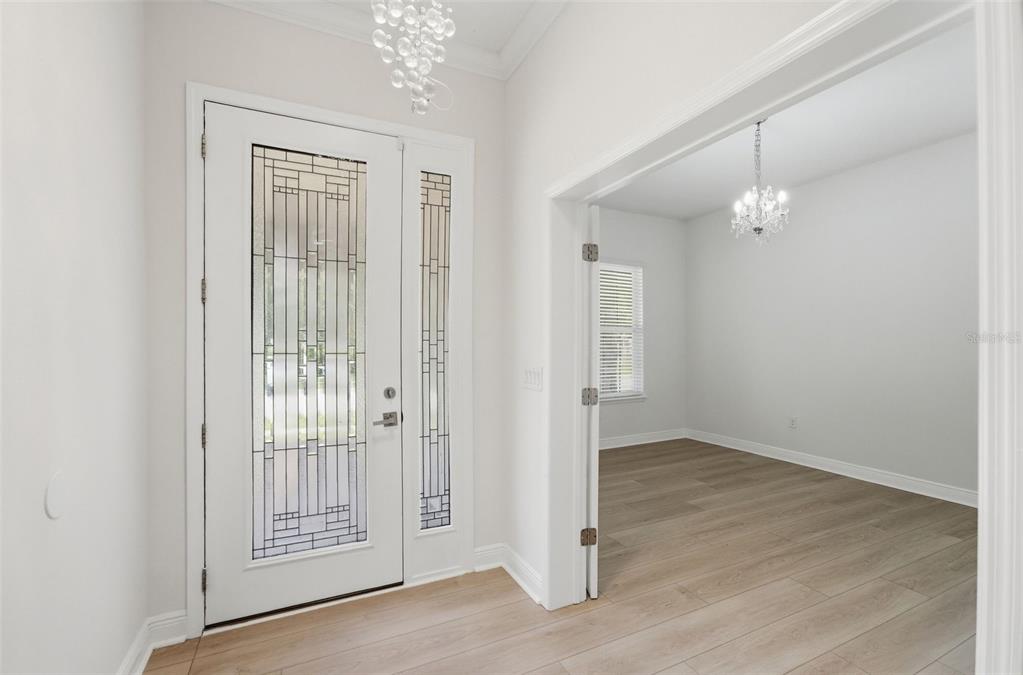An entryway with a glass-paneled white door, light wood flooring, and two crystal chandeliers — 15778 SWEET LIMETTA DRIVE, WINTER GARDEN, FL
