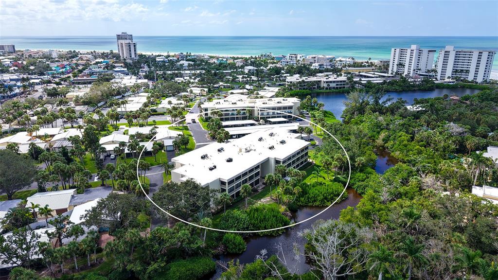 An aerial view shows a multi-story apartment building surrounded by lush greenery and a pond — 225 HOURGLASS WAY UNIT 202, SARASOTA, FL