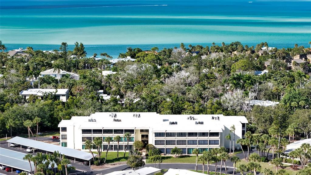 An aerial view shows a multi-story white apartment building surrounded by lush trees near water — 225 HOURGLASS WAY UNIT 202, SARASOTA, FL
