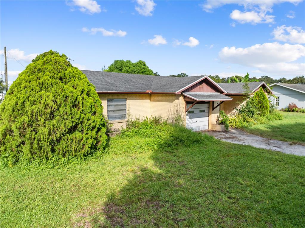 A single-story yellow brick house features a garage, a large evergreen bush, and overgrown landscaping — 740 SHORE DRIVE, KISSIMMEE, FL