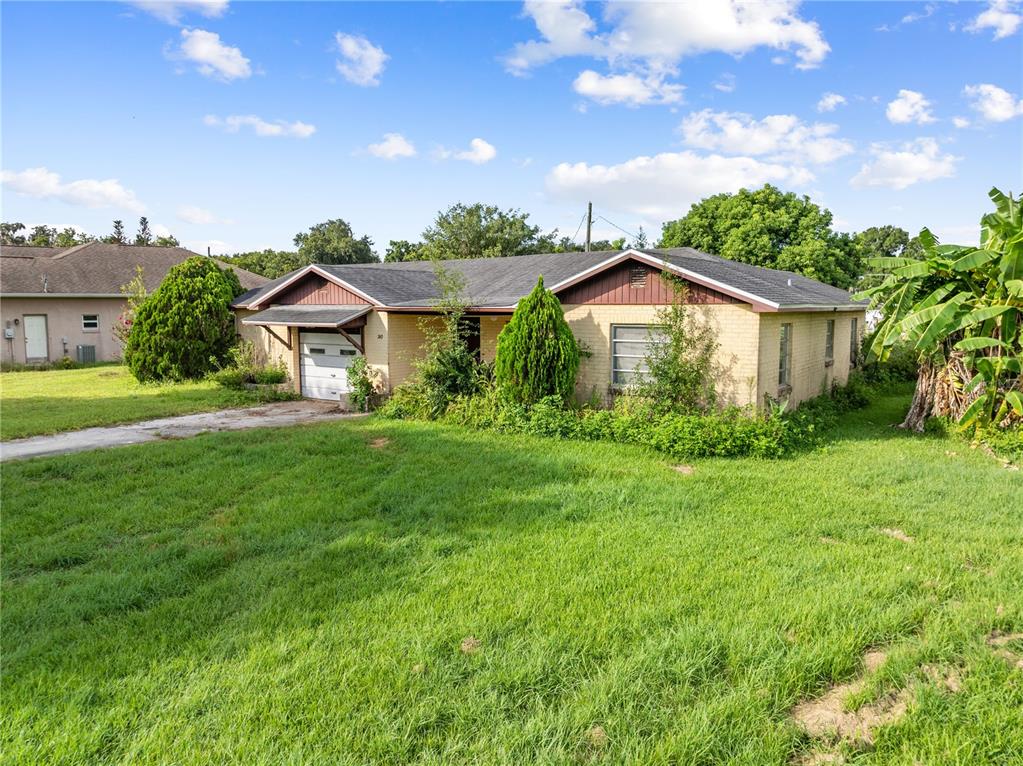 A single-story brick house with a garage and overgrown landscaping under a blue sky — 740 SHORE DRIVE, KISSIMMEE, FL