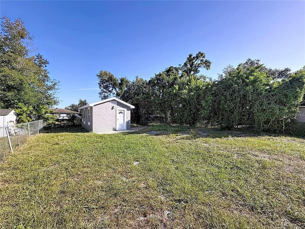 A small, light-colored detached shed sits in a grassy backyard surrounded by dense trees — 2408 E MAGNOLIA STREET, LAKELAND, FL