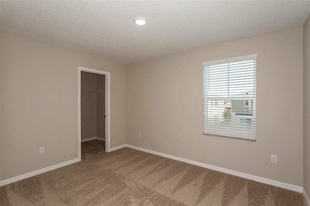 An empty bedroom features beige walls, tan carpeting, a window with blinds, and a walk-in closet — 6051 PRAIRIE RUN, HAINES CITY, FL
