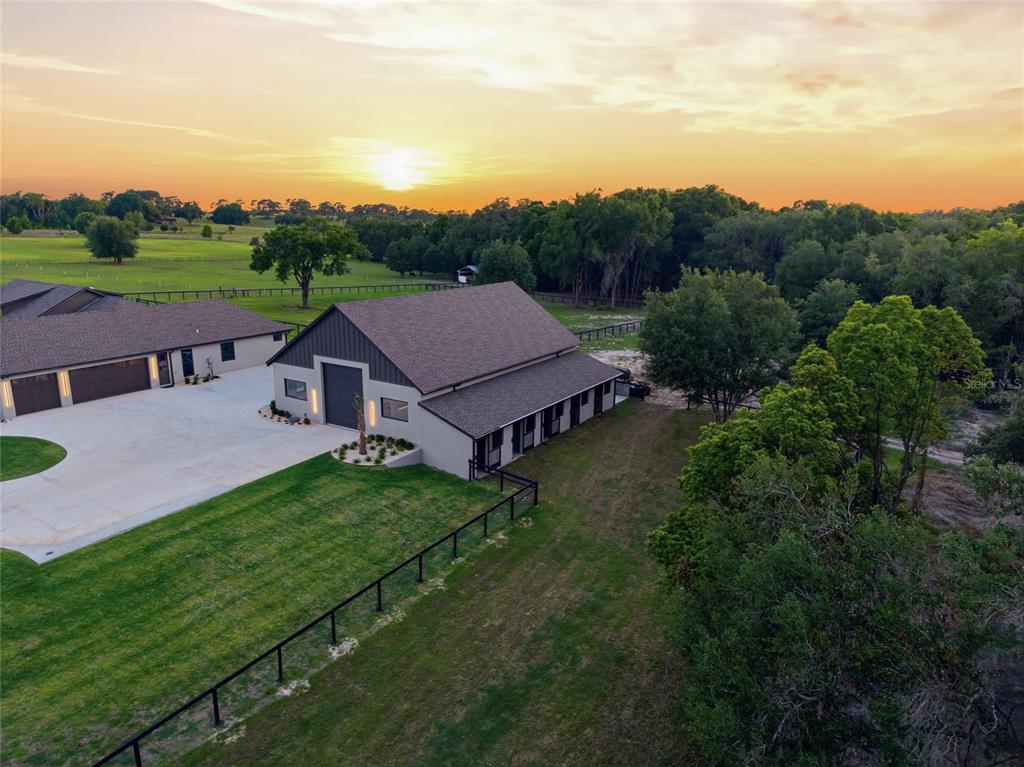 An aerial view shows a modern barn-style building with a concrete driveway at sunset — 3888 SW 140TH AVENUE, OCALA, FL