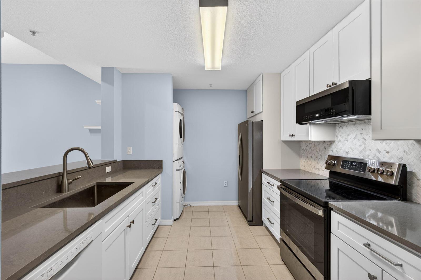 A kitchen with white cabinets, dark countertops, stainless steel appliances, and a stacked laundry unit — 1203 Town Center Drive Drive # 308, Jupiter FL 33458, JUPITER, FL