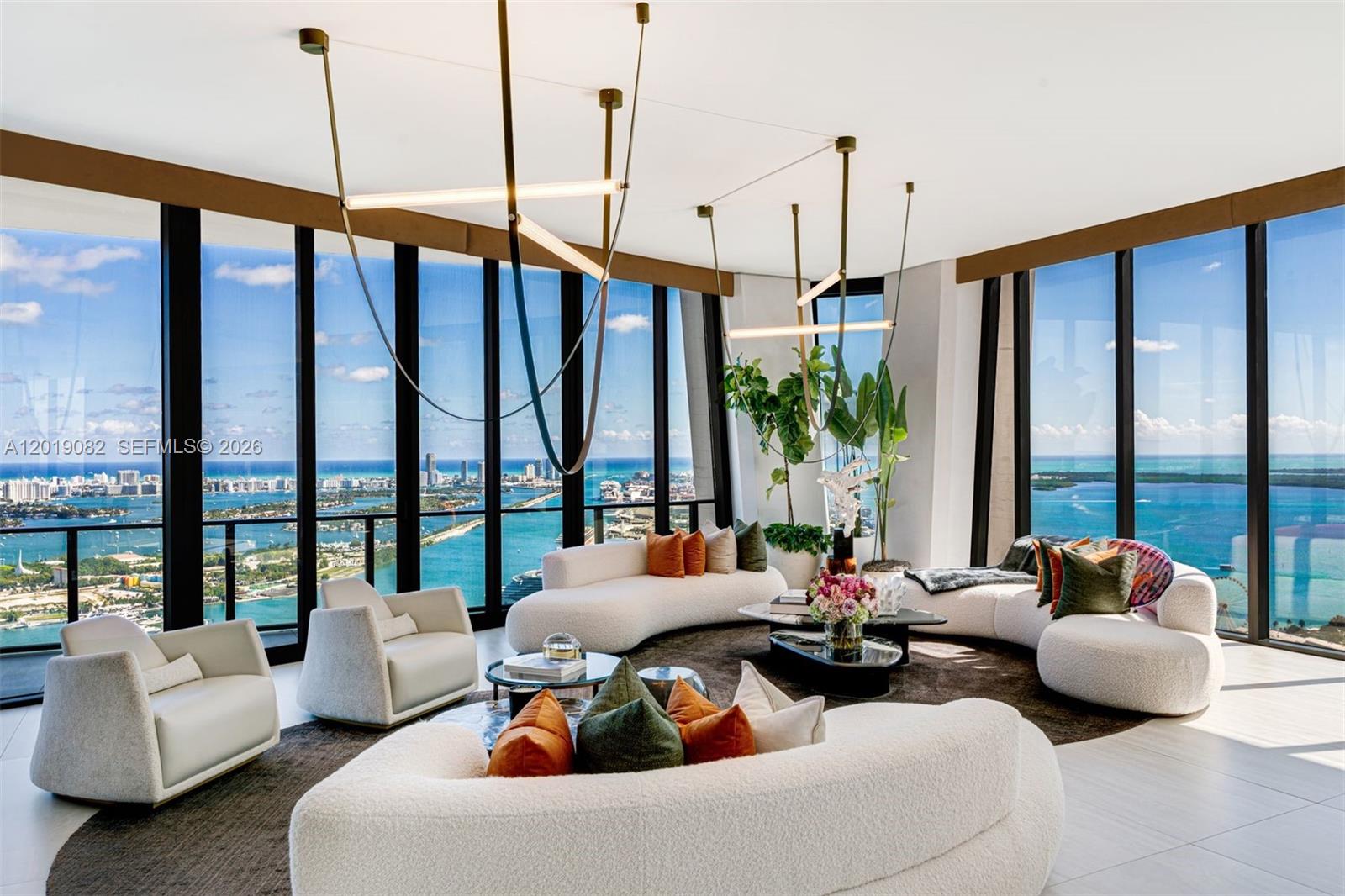 A modern living room features curved white sofas, unique pendant lighting, and floor-to-ceiling ocean views — 1000 Biscayne Blvd # 5101, Miami FL 33132, MIAMI, FL
