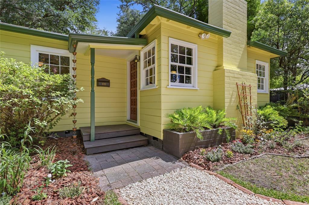 A yellow house exterior features a brick chimney, a small wooden porch, and landscaped gardens — 616 NW 8TH PLACE, GAINESVILLE, FL