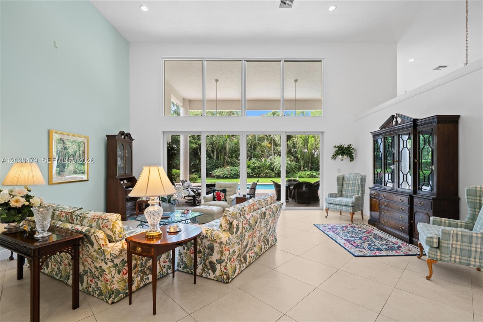 A bright living room features high ceilings, floral furniture, and large windows overlooking a pool — 6020 W Suburban Dr, Pinecrest FL 33156, PINECREST, FL