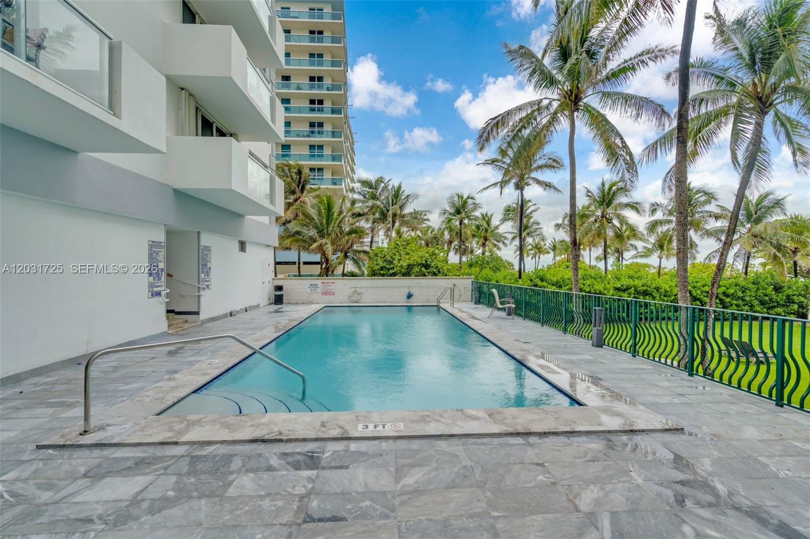 An outdoor swimming pool is surrounded by a tiled deck, palm trees, and a green fence — 9195 Collins Ave # 601, Surfside FL 33154, SURFSIDE, FL