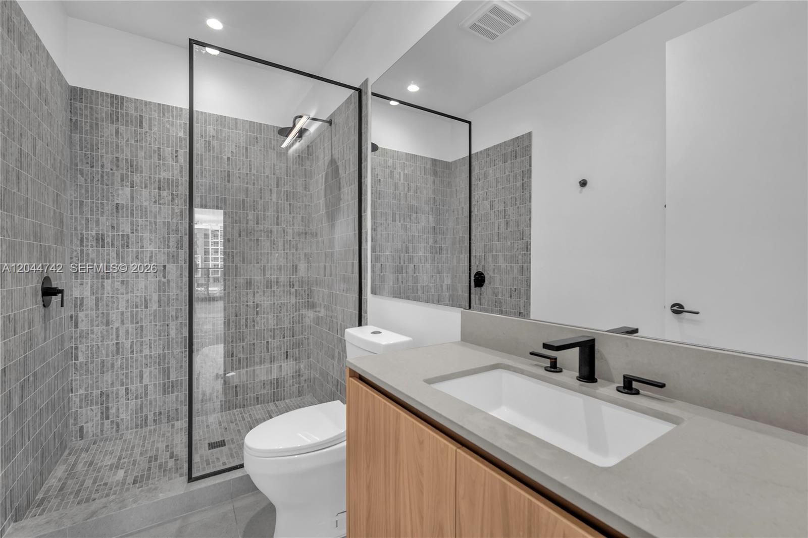 A modern bathroom features a glass-enclosed shower with gray tile, a wood vanity, and mirror — 91 NW 27th St # 201, Miami FL 33127, MIAMI, FL