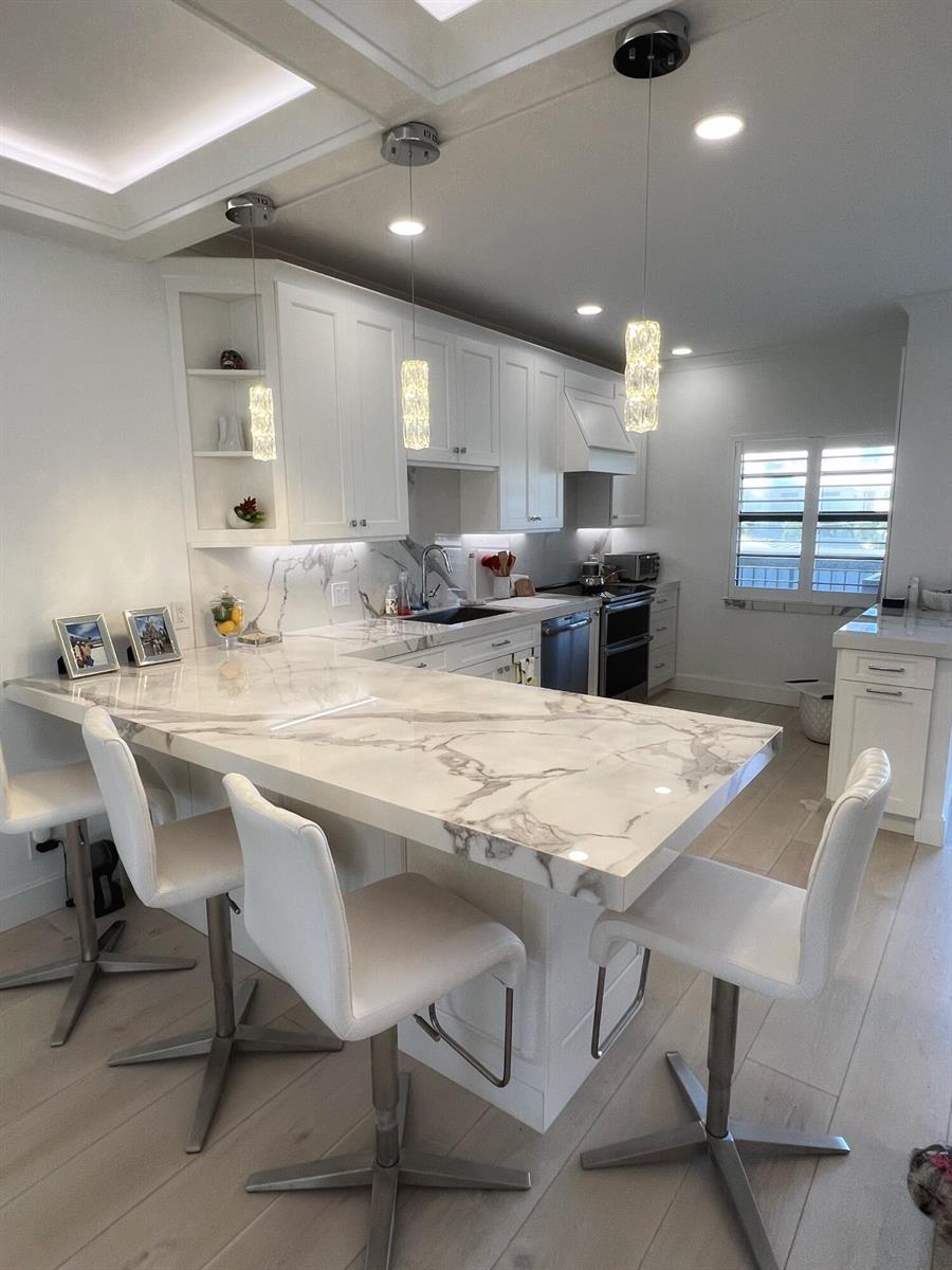 A modern kitchen features white cabinetry, marble countertops, a breakfast bar, and three white stools — 275 S Beach Road # B201, Tequesta FL 33469, TEQUESTA, FL