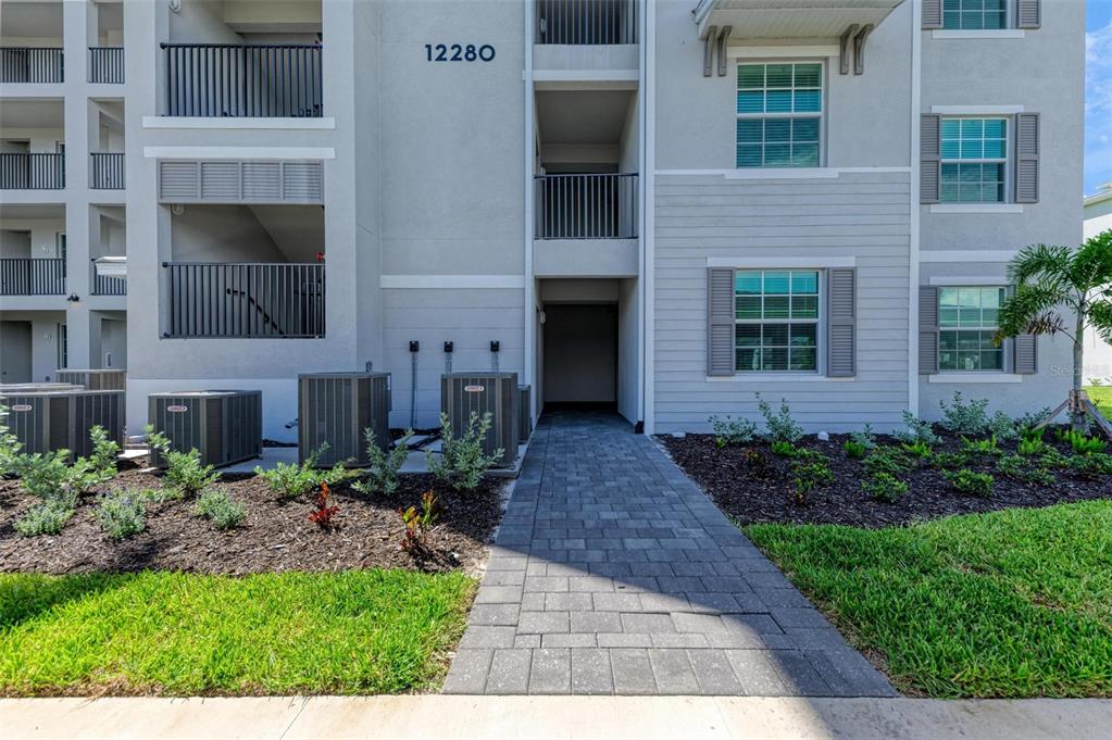 A paved walkway leads to the entrance of a multi-story apartment building with exterior HVAC units — 12280 WELLEN GOLF STREET UNIT 101, VENICE, FL