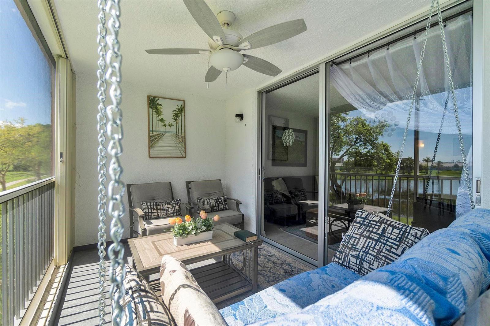 A screened-in balcony features a hanging swing, two armchairs, a coffee table, and ceiling fan — 14575 Bonaire Boulevard # 202, Delray Beach FL 33446, DELRAY BEACH, FL
