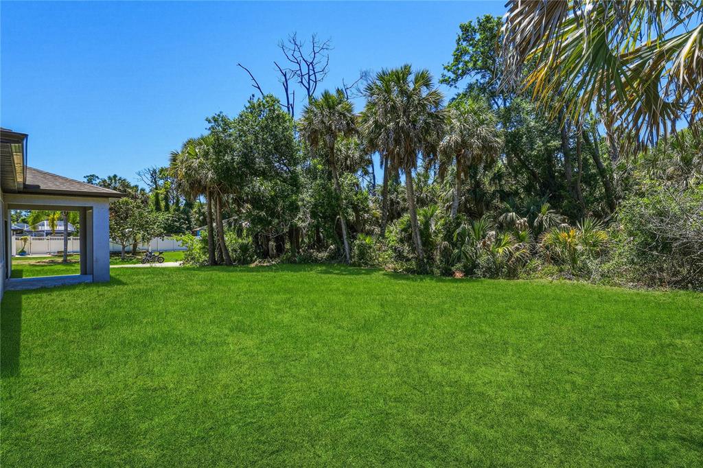 A grassy backyard features a covered patio and dense trees under a clear blue sky — 1459 N CHAMBERLAIN BOULEVARD, NORTH PORT, FL