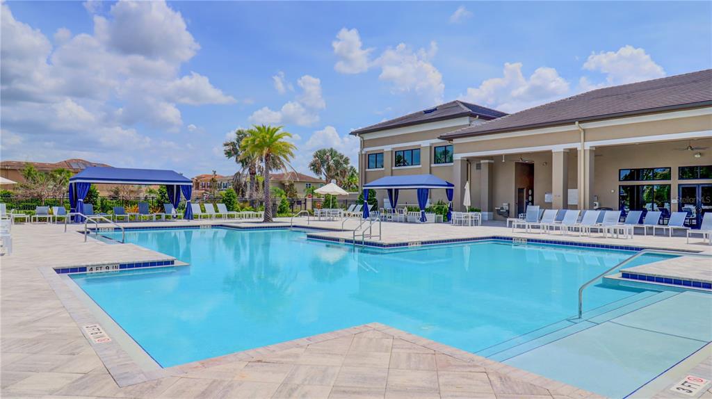 A swimming pool with blue umbrellas and lounge chairs sits beside a large clubhouse building — 3338 GREAT BANYAN WAY, WESLEY CHAPEL, FL