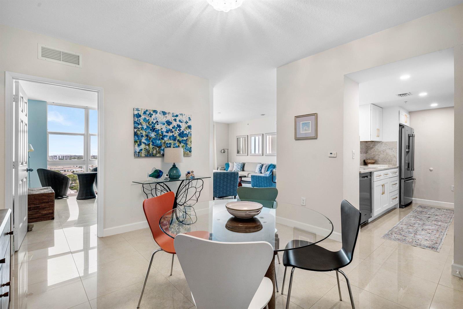 A dining area with a glass table and colorful chairs opens to a kitchen and balcony — 625 Casa Loma Boulevard # 1408, Boynton Beach FL 33435, BOYNTON BEACH, FL