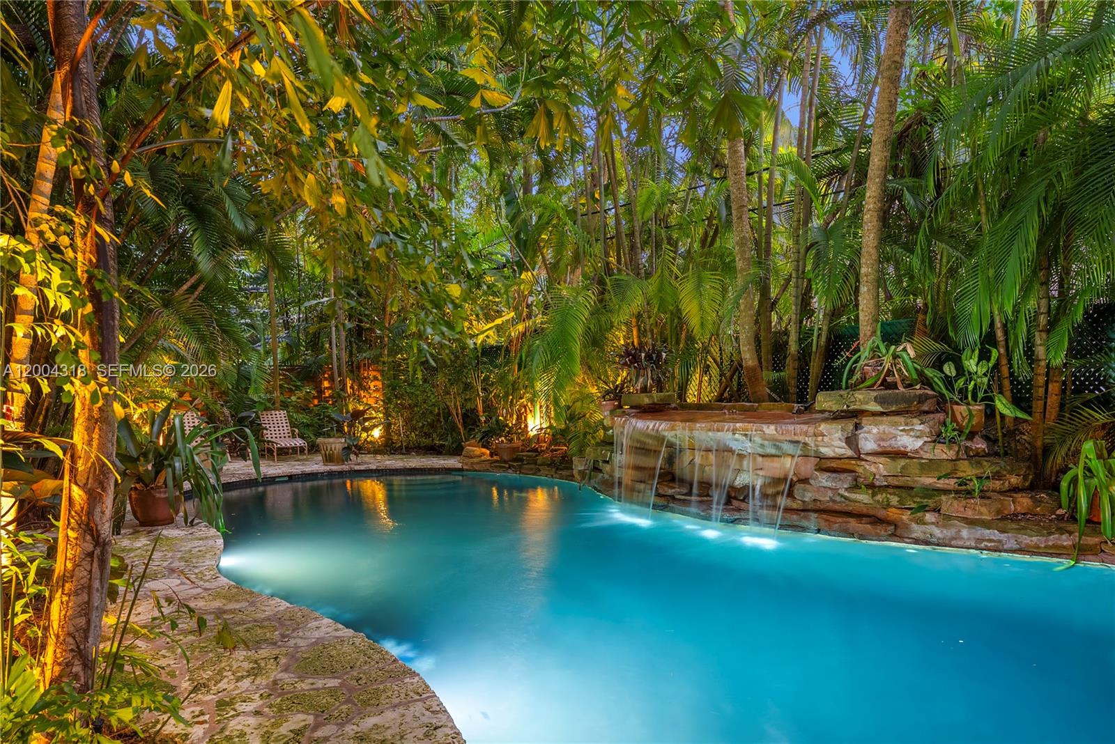 A swimming pool with a stone waterfall feature is surrounded by lush tropical trees — 3584 Saint Gaudens Rd, Miami FL 33133, MIAMI, FL