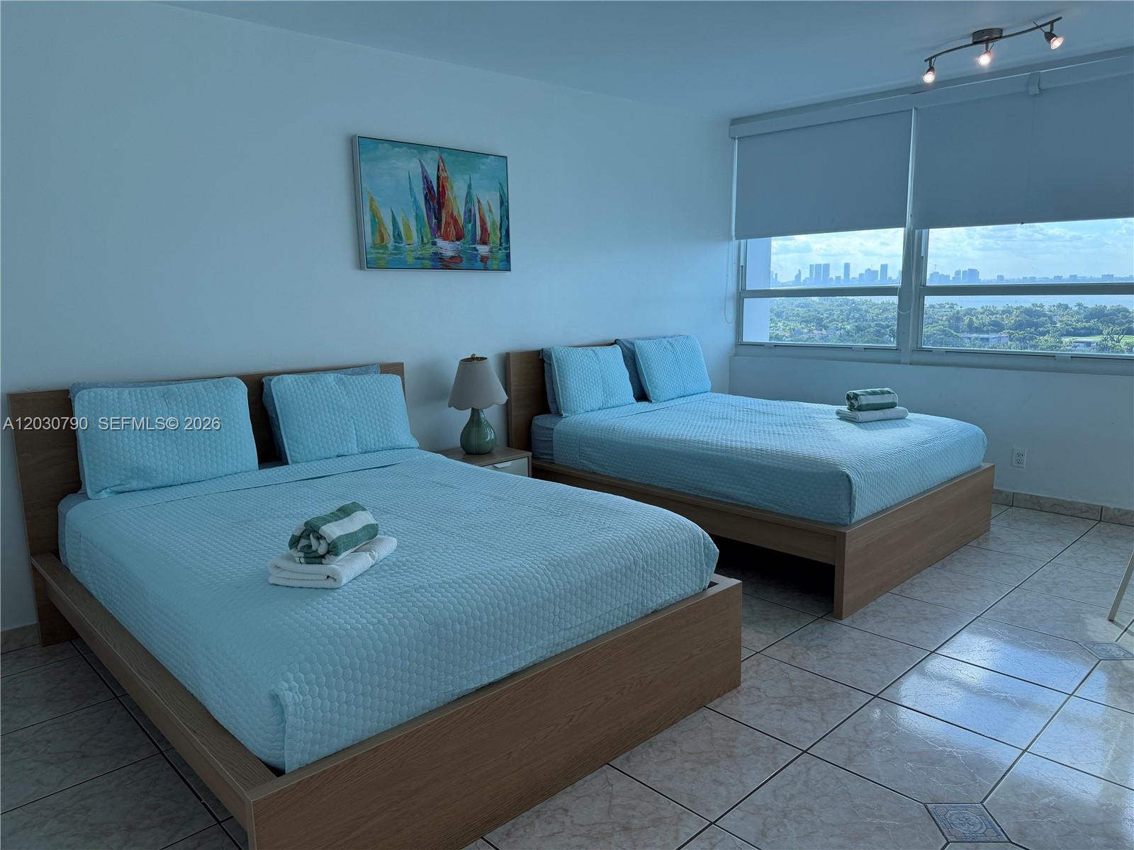 A bedroom features two beds with light blue bedding, tile floors, and a city view — 5445 Collins Ave # 1506, Miami Beach FL 33140, MIAMI BEACH, FL
