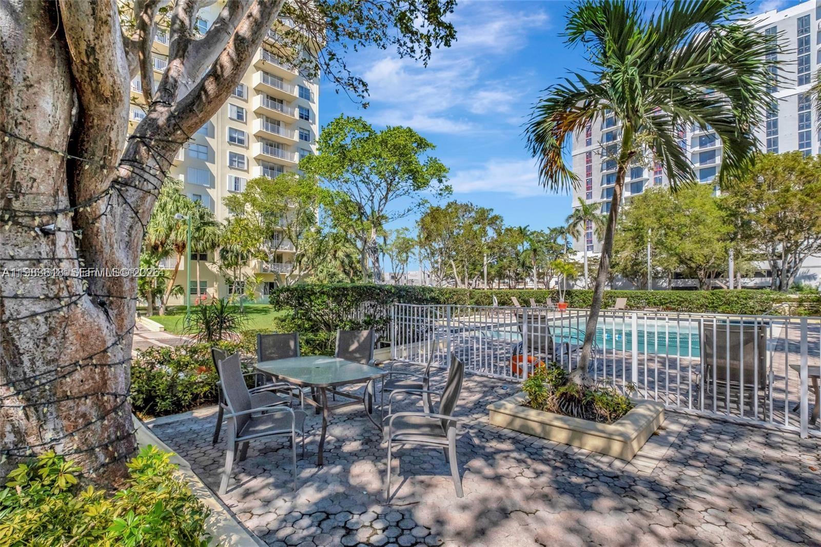 A patio with a glass-top table and chairs sits beside a swimming pool and buildings — 11111 Biscayne Blvd # 8F, Miami FL 33181, MIAMI, FL