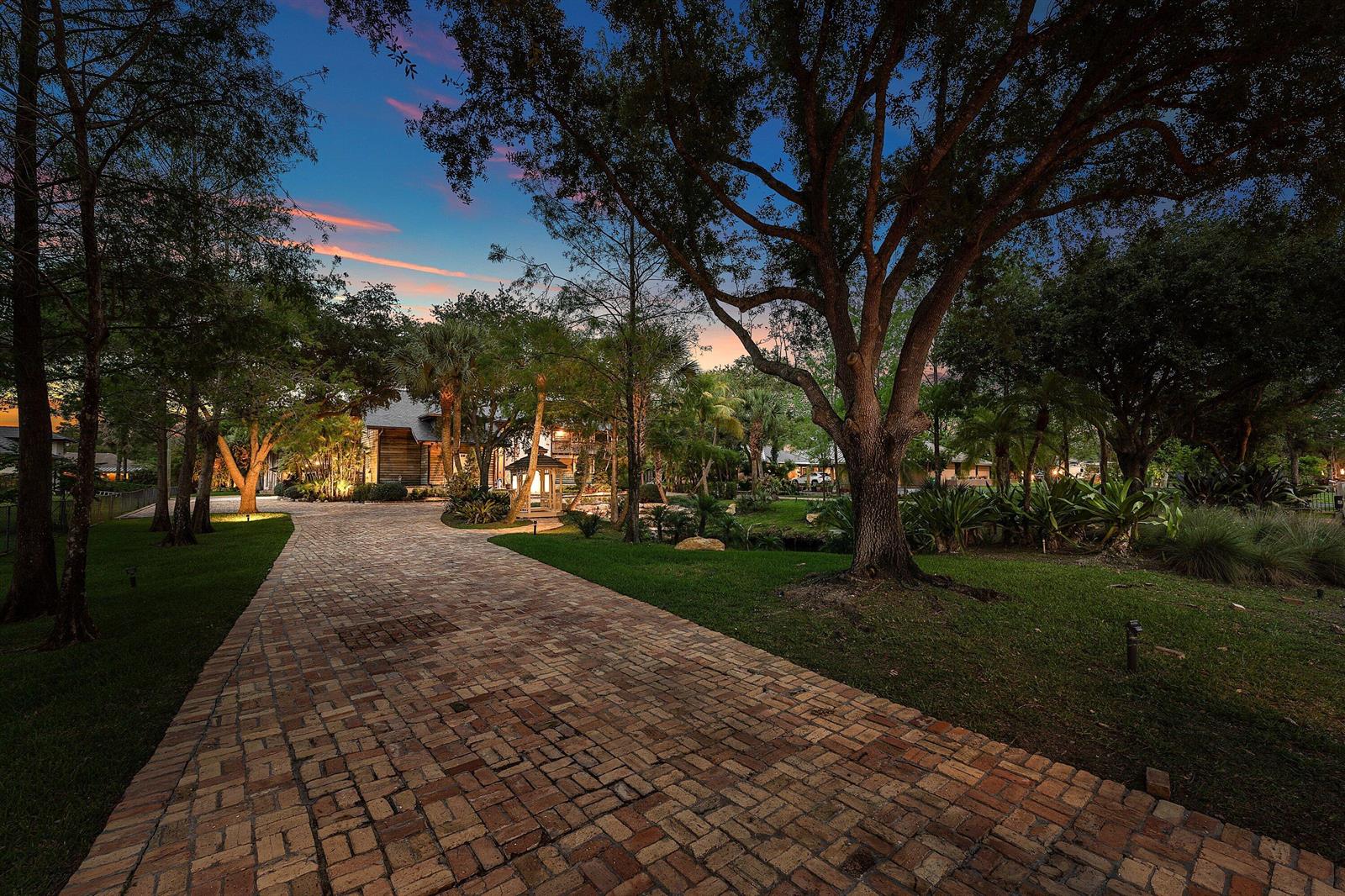 A brick driveway leads toward a house surrounded by trees and lush landscaping at dusk — 6002 NW 74th Terrace, Parkland FL 33067, PARKLAND, FL