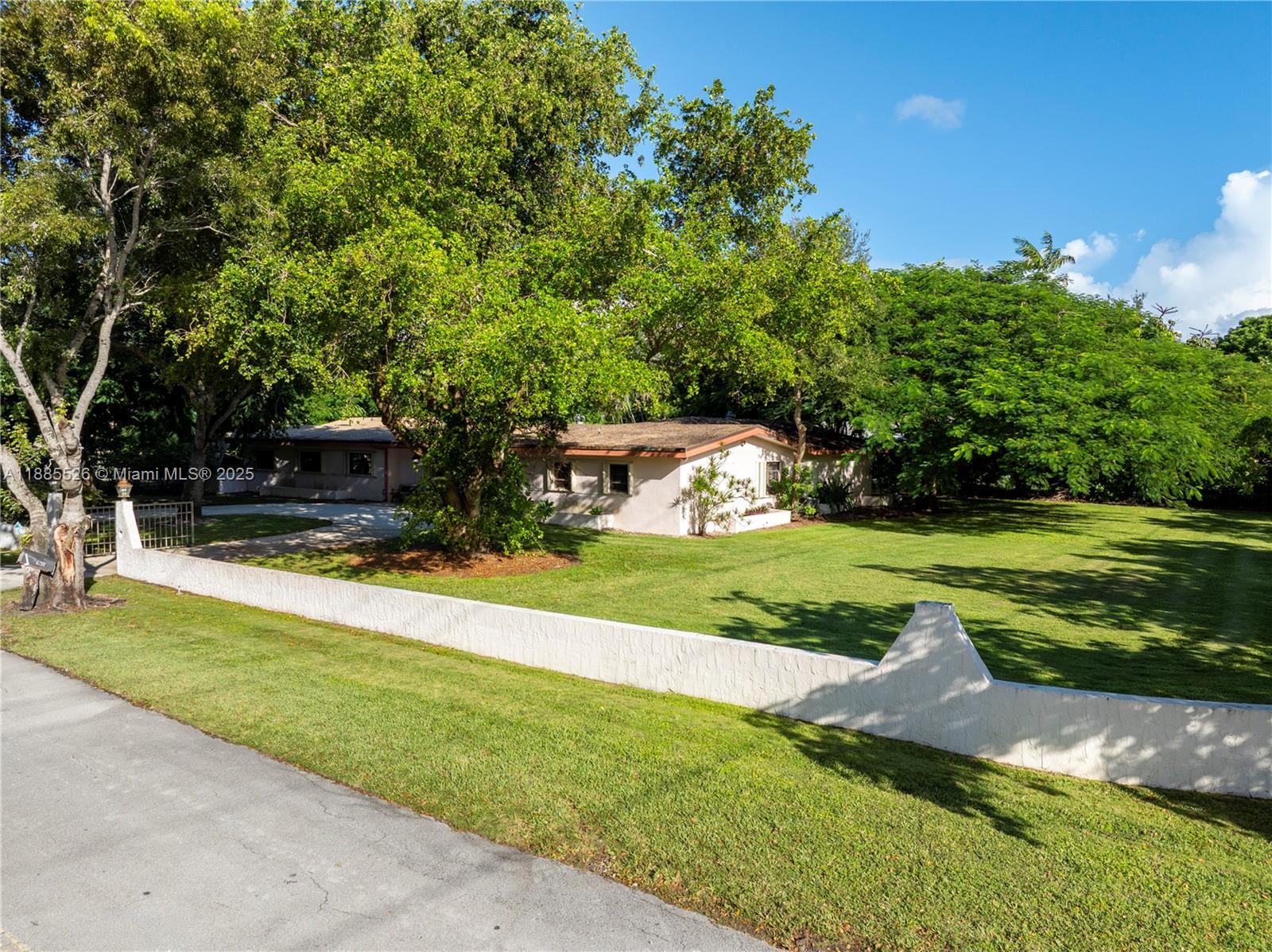 A single-story house with a white wall sits behind a large lawn and trees — 6201 SW 114th St, Pinecrest FL 33156, PINECREST, FL