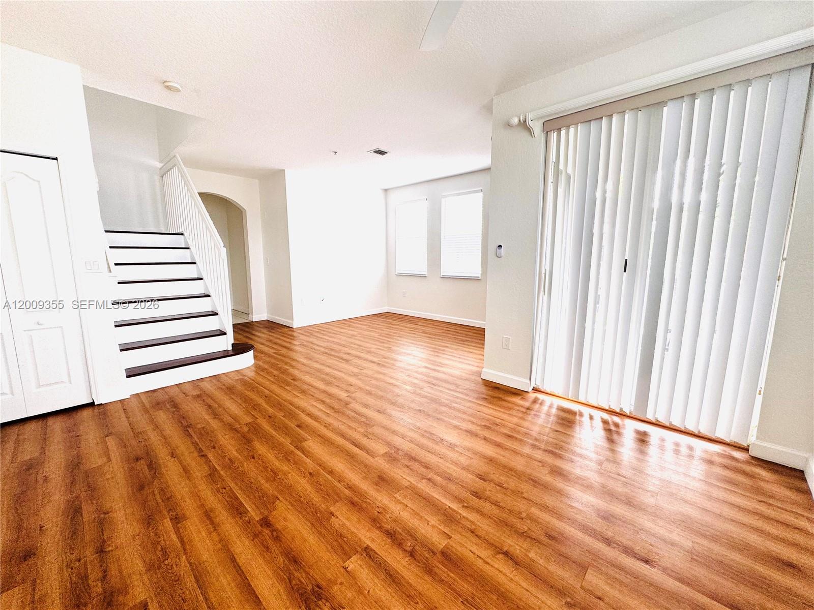 An empty living room features wood-look flooring, a staircase, and vertical blinds on sliding doors — 4435 SW 160th Ave # 214, Miramar FL 33027, MIRAMAR, FL