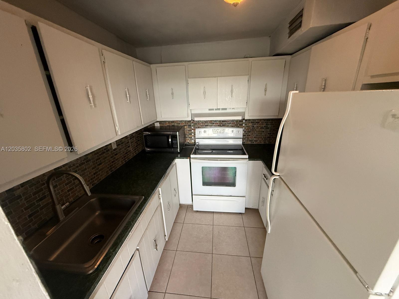 A galley kitchen features white cabinets, dark countertops, a tiled backsplash, and white appliances — 6675 W 4th Ave # 402, Hialeah FL 33012, HIALEAH, FL