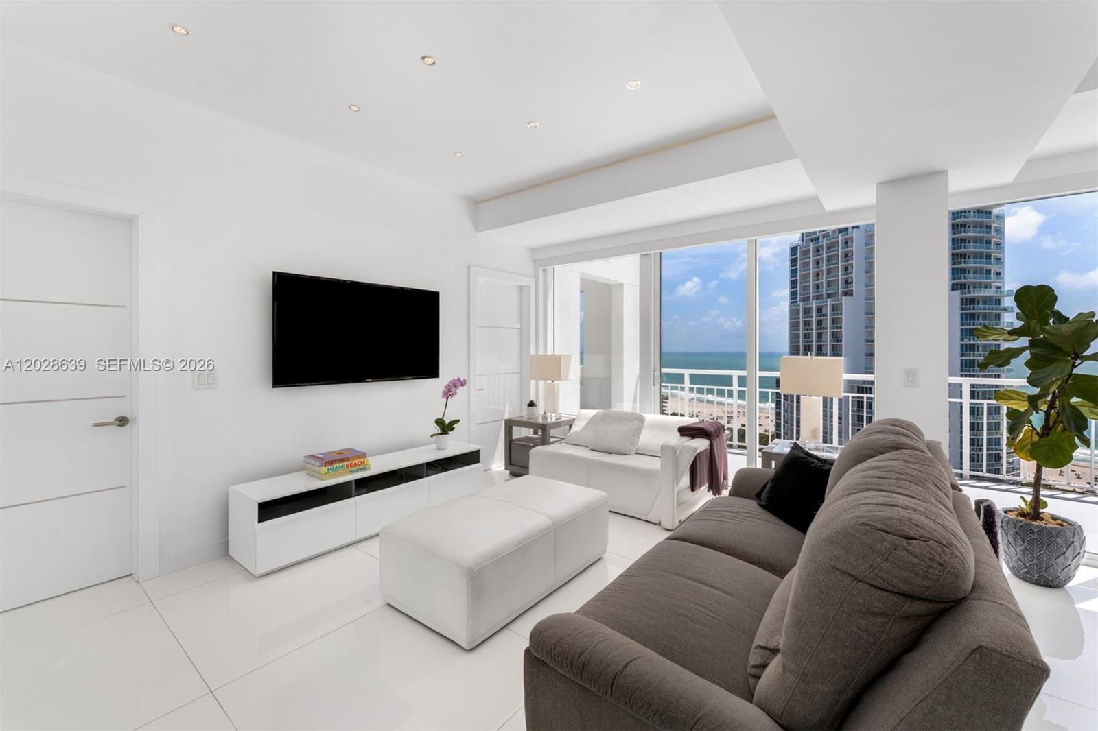 A modern living room features a grey sofa, white furniture, and floor-to-ceiling ocean view windows — 400 S Pointe Dr, Miami Beach FL 33139, MIAMI BEACH, FL