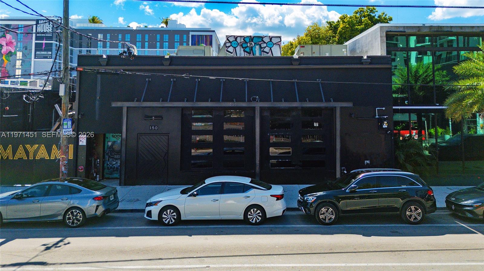 A black commercial building with large glass garage doors sits behind a row of parked cars — 150 NW 24th St, Miami FL 33127, MIAMI, FL