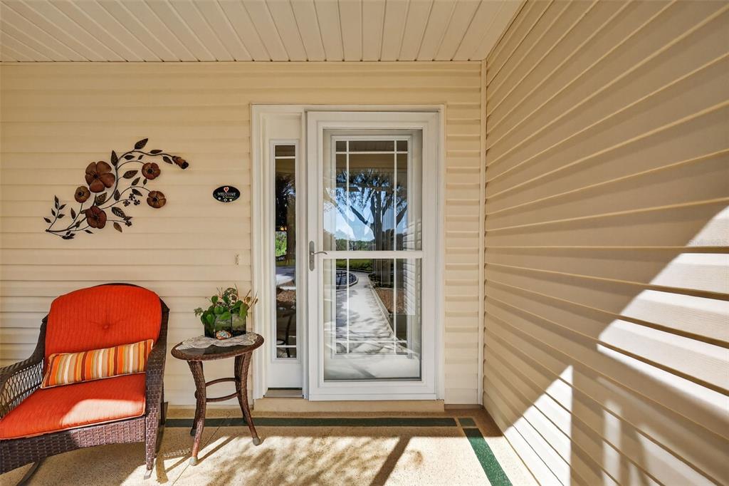 A covered porch features beige vinyl siding, a glass storm door, and an orange wicker chair — 416 CARRERA DRIVE, THE VILLAGES, FL