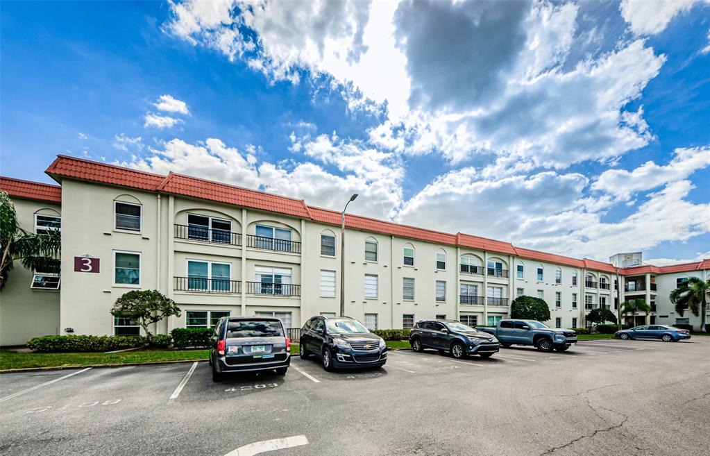 A multi-story apartment building with a red tile roof and a parking lot in front — 2700 BAYSHORE BOULEVARD UNIT 3208, DUNEDIN, FL