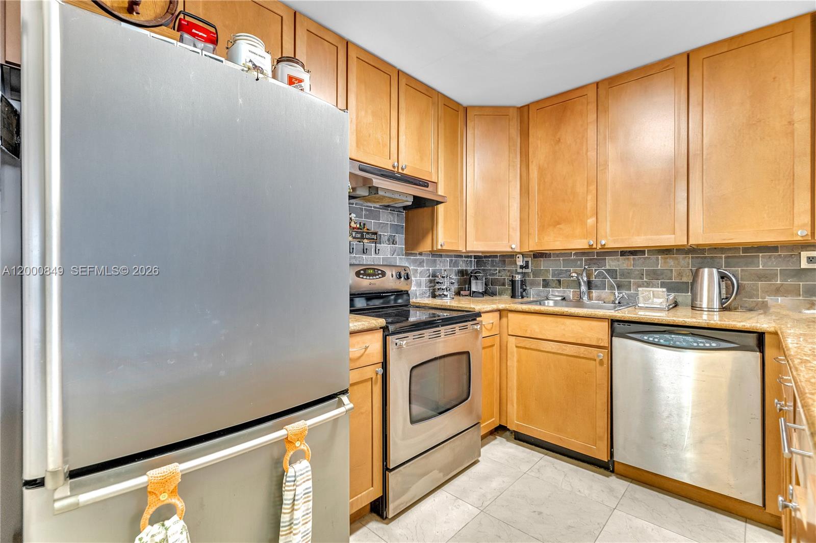 A kitchen features light wood cabinets, stainless steel appliances, and a gray stone backsplash — 600 Three Islands Blvd # 618, Hallandale Beach FL 33009, HALLANDALE BEACH, FL