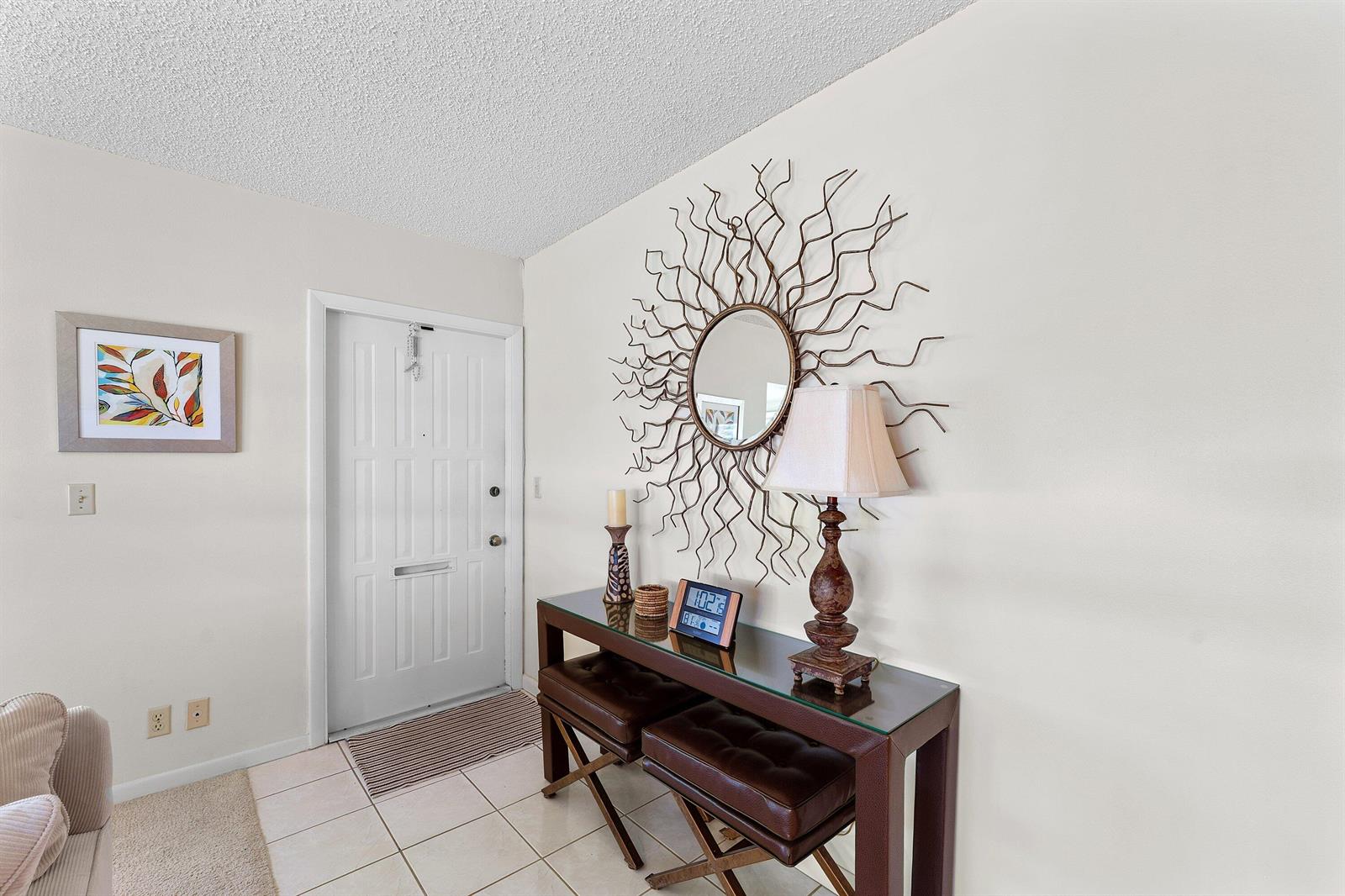 An entryway features a glass-topped console table, two leather stools, and a sunburst wall mirror — 1210 S Drive Circle # D, Delray Beach FL 33445, DELRAY BEACH, FL