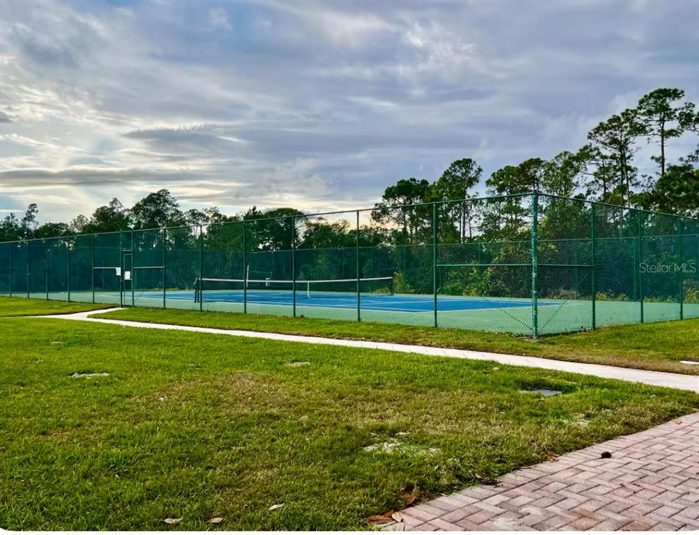 A fenced-in outdoor tennis court sits on a grassy lot under a cloudy sky — 1820 CORIANDER DRIVE, POINCIANA, FL