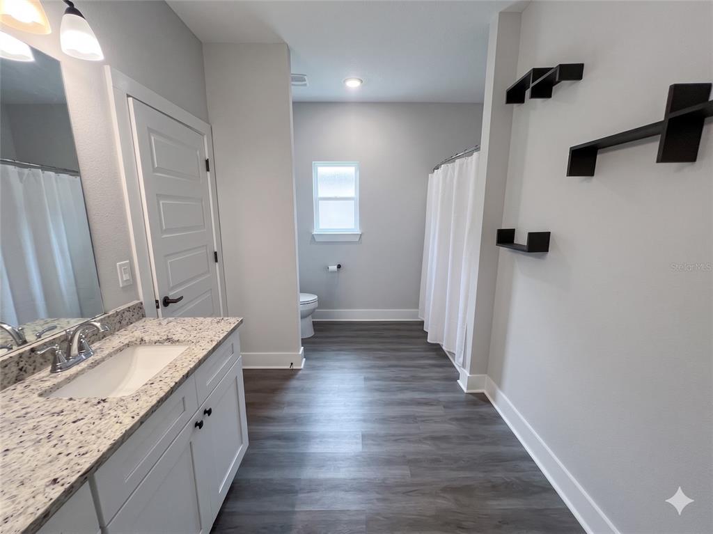 A bathroom features a granite vanity, dark wood-look flooring, a shower curtain, and wall shelves — 3044 N 7TH AVENUE, MILTON, FL
